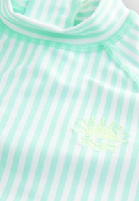 Haut de bain rayé vert clair et blanc avec un col haut et des manches courtes, présentant un motif de soleil brodé jaune à l'avant.