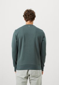 Mies, jolla on kiharat hiukset, pukeutuneena tummanvihreään pitkähihattomaan paita ja vaaleanharmaisiin housuihin, seisoo selkä kameraan päin yksinkertaista taustaa vasten.