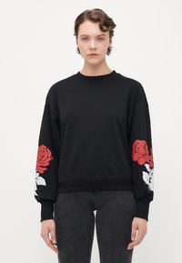 Ungdom med kort krøllete hår iført svart hettegenser med røde og hvite roseutskrifter på begge ermer, kombinert med mørke jeans.