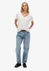 Mujer de pie con las manos detrás de la espalda, vistiendo una camiseta blanca de cuello en V, jeans azul claro con un cinturón marrón y mocasines negros.