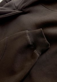 Felpa con cappuccio marrone caratterizzata da una texture morbida, polsini a coste e una tasca a marsupio. Il tessuto ha una finitura liscia con dettagli cuciti.