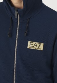 Giacca zip-up blu navy con tessuto testurizzato, colletto alto e una patch con logo dorato sul lato sinistro del petto. Presenta lacci neri e una zip argentata.