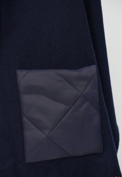 Îmbrăcăminte din lână bleu marin, cu un buzunar din material pătrățos, matlasat, cu un model în diamant, care sporește textura și funcționalitatea.
