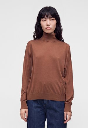 Vrouw met lang zwart haar, een bruine coltrui en blauwe spijkerbroek, staand tegen een eenvoudige lichte achtergrond.