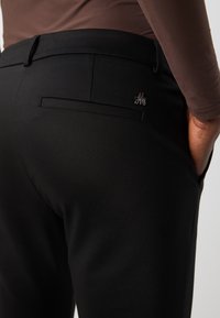 Černé kalhoty s hladkým povrchem, které mají jednu zadní kapsu a malý kovový logo prvek poblíž kapsy.