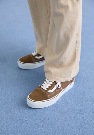 Hnědé a bílé tenisky na šněrování nošené s béžovými kalhotami širokých nohavic na světle modrém koberci.