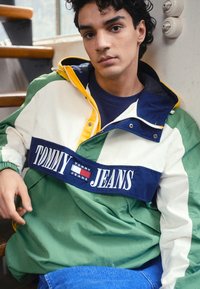 Krāsains vēja aizsargs zaļā, baltā un tumši zilā krāsā ar rāvējslēdzēja apkakli, elastīgām piedurknēm un uzkrītošu "TOMMY JEANS" logo krūšu daļ�ā.