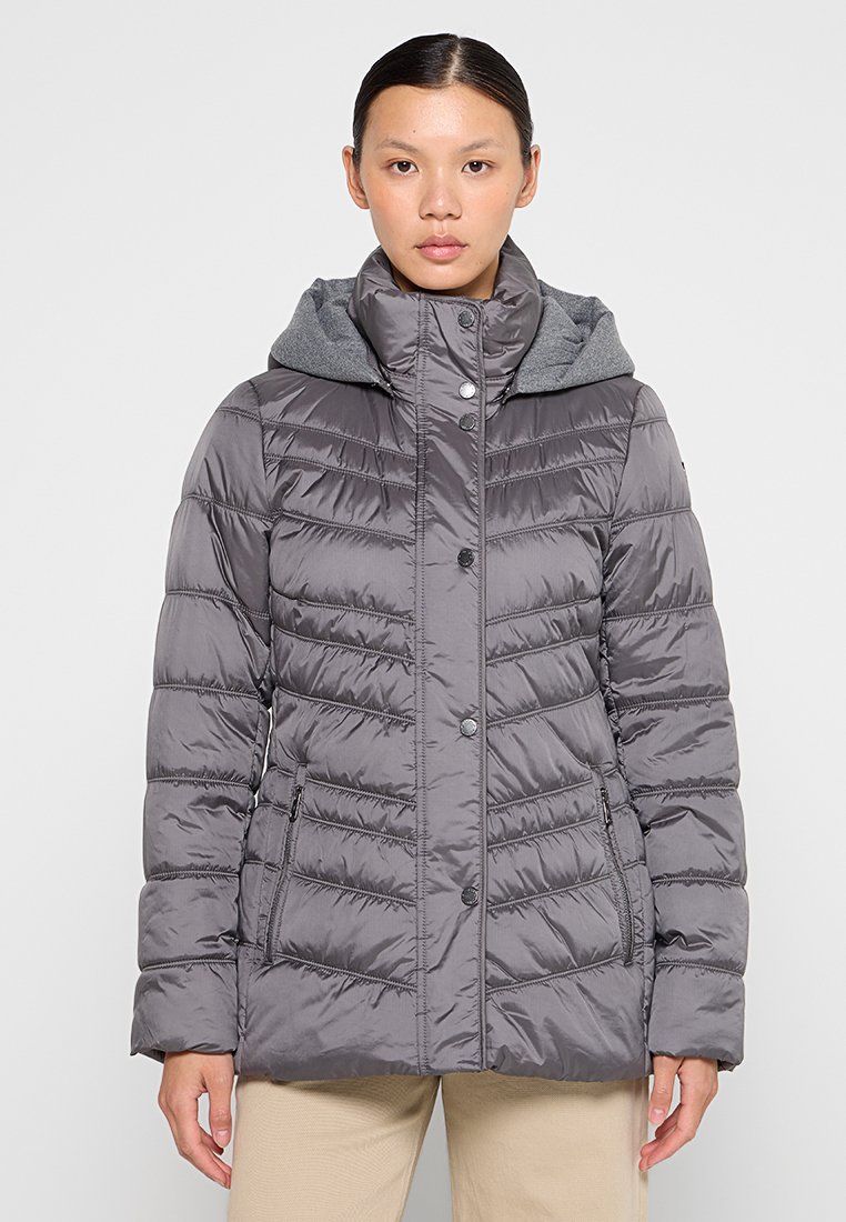 Kobieta ubrana w szary pikowany zimowy płaszcz z wysokim kołnierzem, z zatrzaskami, kieszeniami z boku na zamek błyskawiczny oraz z przymocowanym kapturem na szarym wewnętrznym elemencie.