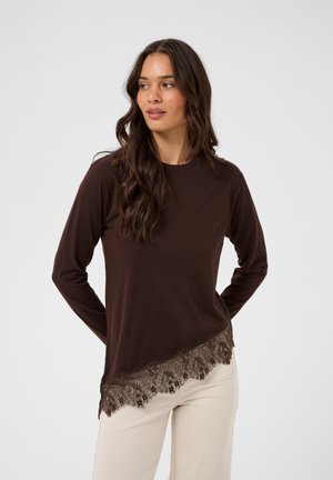 Femme aux longs cheveux bruns portant un haut à manches longues marron foncé avec une bordure en dentelle et un pantalon crème, debout devant un fond clair uni.