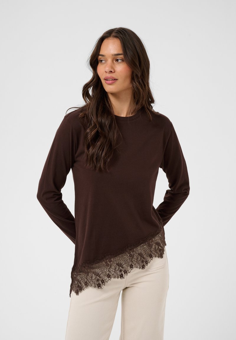 Femme aux longs cheveux bruns portant un haut à manches longues marron foncé avec une bordure en dentelle et un pantalon crème, debout devant un fond clair uni.