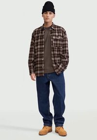 Jaunas vyras dėvi juodą kepuraitę, rudą languotą marškinėlius ant pilko marškinio, mėlynus džinsus ir šviesiai rudas darbo batus, stovintis prieš vienodą foną.