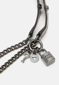Metal kæderem med læderdetaljer, der har dekorative vedhæng: hængelås, spørgsmålstegn og trekant, alle med sort finish.