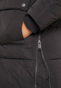 Ruka v kapse černé péřové bundy s viditelným stříbrným zipem a patentkem na látkové klopě nad kapsou.