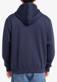 Felpa blu navy con cappuccio, realizzata in tessuto morbido. Presenta polsini e orlo a costine, con un piccolo logo dettagliato sulla manica sinistra.