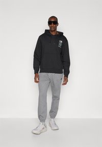 Sweatshirt preto com capuz e bolsa frontal, detalhe do logo no peito, acompanhado de calças de moletom cinzentas, ténis brancos e óculos de sol grandes na cor preta.