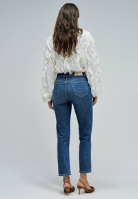 Mujer con cabello largo castaño que lleva una blusa blanca con volantes, jeans azules y sandalias de tacón marrones, de pie con la espalda hacia la cámara.