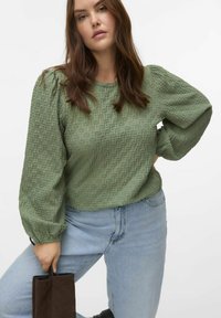 Maglione verde con texture, maniche a sbuffo, scollo rotondo e motivo sottile, abbinato a jeans azzurri chiari e una borsa marrone.