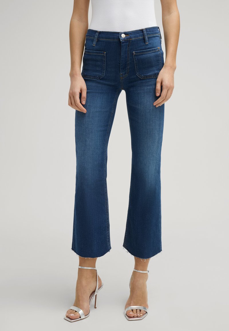 Jeans z wysokim stanem, ciemnoniebieskie, z rozszerzonym dołem. Posiadają dwie przednie kieszenie oraz zapięcie na guzik. Gładka tekstura i brak widocznych wzorów.