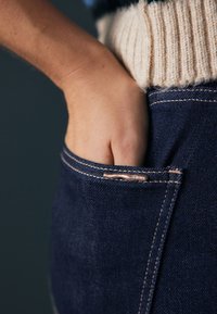 Mano insertada en el bolsillo delantero de unos vaqueros azul oscuro, combinados con un suéter beige de punto.