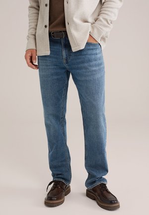 Man in blauwe rechtvallende jeans, donkerbruine riem, beige cardigan over een bruine shirt en donkerbruine veterschoenen, staand op een lichte achtergrond.