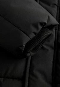 Detail čiernej puffrovanej bundy so hladkou matnou tkaninou, hrubou vycpanou rukávom a akcentom z rebrového pleteného manžetu.