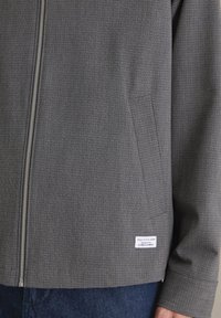 Szara kurtka zapinana na zamek, wykonana z teksturowanej tkaniny z subtelnym geometrycznym wzorem, wyposażona w boczne kieszenie i metkę marki przy dolnym brzegu.