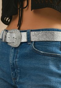 Sølvglitrende bælte med en stor rhinstenbesat spænde, parret med blå denim jeans, der fremhæver tekstur og metalliske accenter.