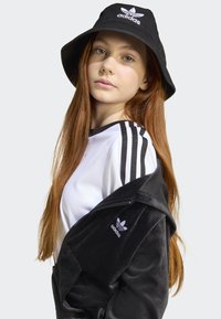 Black velvet jacket with embroidered Adidas logo, white athletic shirt featuring black stripes, and a black bucket hat with logo. Long red hair.