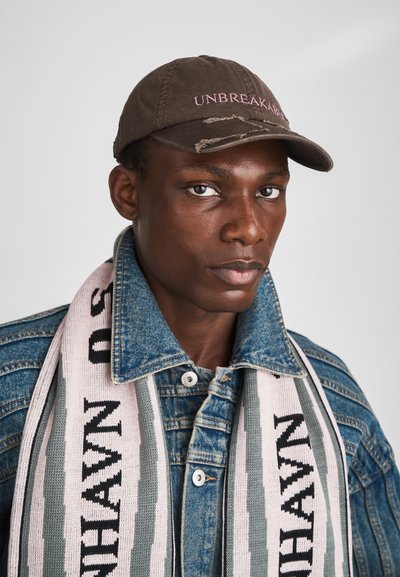 Gorra de béisbol marrón con "UNBREAKABLE" en texto rosa. Chaqueta de mezclilla con una textura ligera. Bufanda de punto a rayas en rosa y verde con texto.