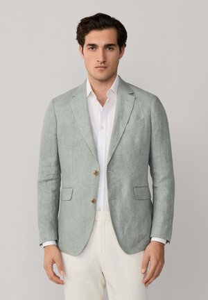 Homme portant un blazer gris clair sur une chemise blanche et un pantalon crème, debout devant un fond uni gris clair.