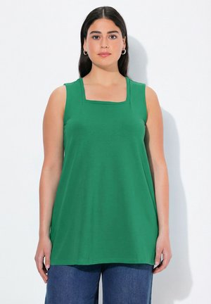 Vrouw met lang donker haar, die hoepeloorringen draagt, een mouwloos groen topje met vierkante halslijn en een blauwe spijkerbroek, staand tegen een witte achtergrond.