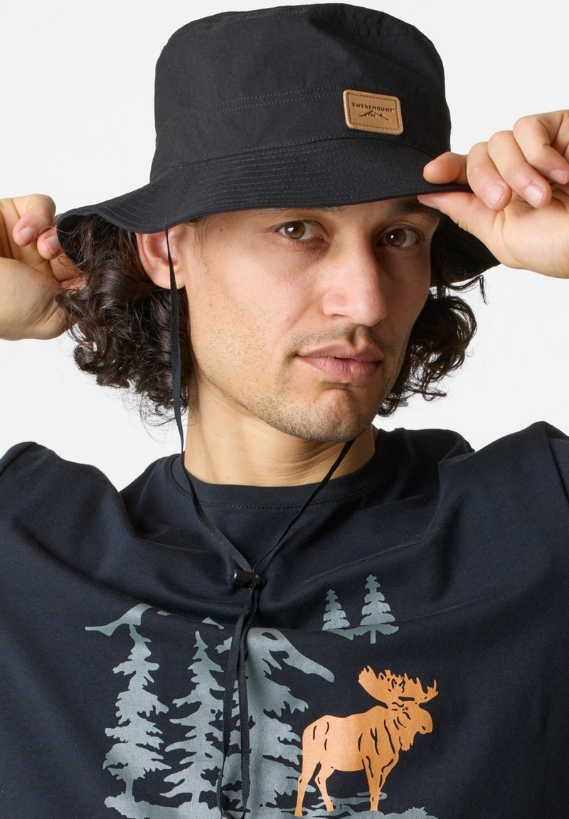 Black bucket hat made of lightweight fabric, featuring a leather tag and an adjustable chin strap for secure wear. Textured surface.