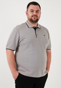 Szürke rövidujjú polo ing, fekete gallérral, kétgombos gombolású, kontrasztos fekete díszítéssel az ujjak mentén. Diszkrét márkájú logó a mellkasán.