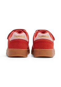 Czerwone zamszowe trampki z gumową podeszwą, z różowym panelem na pięcie i złotym logo "hummel". Zapięcie na rzep zapewniające pewne dopasowanie.