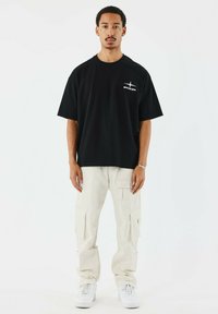 Camiseta negra de gran tamaño con gráfico blanco en el pecho, combinada con pantalones cargo de color beige claro que presentan múltiples bolsillos y un ajuste holgado. Zapatillas blancas.