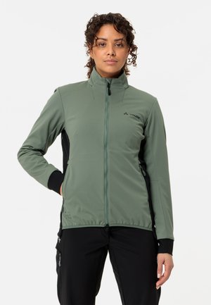 Frau mit lockigem Haar, die eine grüne Jacke mit Reißverschluss und schwarze Hose trägt, steht mit einer Hand in der Tasche vor einem schlichten weißen Hintergrund.