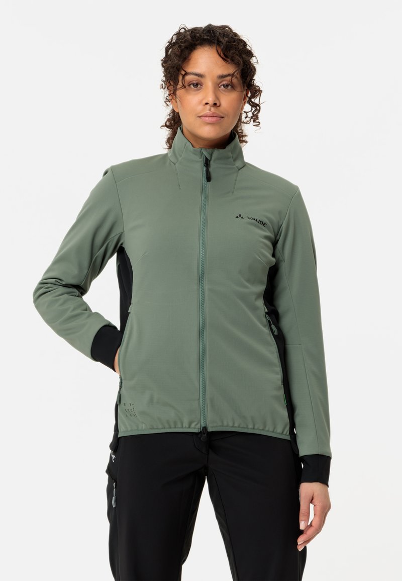 Femme aux cheveux bouclés portant une veste verte zippée et un pantalon noir, debout avec une main dans la poche sur un fond blanc uni.