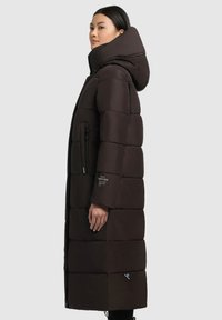 Manteau rembourré long marron avec un col haut et une capuche, doté d'une poche zippée sur le côté et d'un détail de logo sur la manche. Texture lisse.