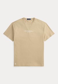 Μπεζ κοντομάνικη μπλούζα με το κείμενο "Polo Ralph Lauren New York" στο στήθος και μικρό λευκό λογότυπο Polo κοντά στο κάτω τελείωμα.