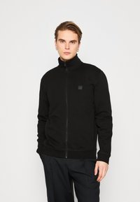 Black zip-up jacket with high collar, side pockets, and a small logo patch on the chest. Soft fabric and relaxed fit. Paired with dark trousers.