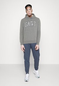 Felpa grigia con logo "GANT", abbinata a pantaloni della tuta blu scuro e sneakers bianche. Il tessuto è morbido e la vestibilità complessiva è rilassata.