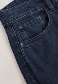 Tamsiai mėlyni džinsai iš džinsinio audinio su išblukusia tekstūra, matomi priekiniai kišenės ir metaliniai vinukai. Švarus siuvimas ir tvirtas juosmuo.