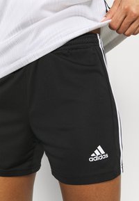 Persona ajustándose una camiseta deportiva blanca, llevando pantalones cortos negros de Adidas con rayas blancas y el logo en la pierna izquierda, contra un fondo liso.