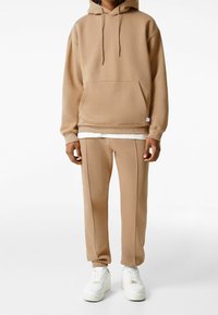 Beige Kapuzenpullover mit Fronttasche und verstellbarem Kordelzug, kombiniert mit passenden beigen Hosen mit seitlichen Nähten. Weiße Sneakers.