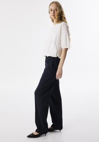 Witte blouse met korte mouwen en een zak, gecombineerd met marineblauwe wijde broeken. Model draagt zwarte puntige ballerina's. Eenvoudig en minimalistisch ontwerp.