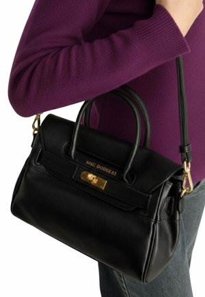 Sac à main Mac Douglas en cuir noir avec fermoir doré et doubles poignées, porté à l'épaule par une personne en pull violet et jean gris.