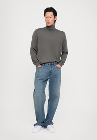 Pulover gri cu guler înalt, croi lejer, asortat cu blugi din denim albastru și adidași albi. Design simplu, material texturat, stil casual.