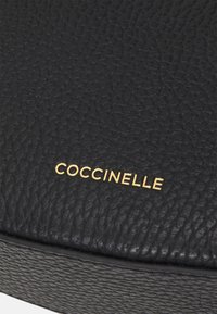 Must mustri nahkakott, millel on sile tekstuur ja millel on kullavärvi "COCCINELLE" logo rõhutatud pinnale.