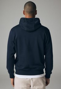Sudadera azul marino hecha de un tejido suave, con capucha, puños y dobladillo de canalé. Mostrada desde atrás, con una camiseta blanca visible por debajo.