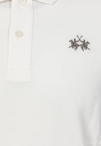 Bílé polo tričko s texturovanou vazbou, s dvouknoflíkovou légou a vyšitým logem dvou jezdců se zkříženými pálkami.